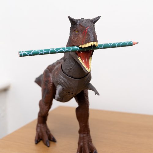 A toy dinosaur holding a green pencil with a dinosaur skin design pencil in its mouth